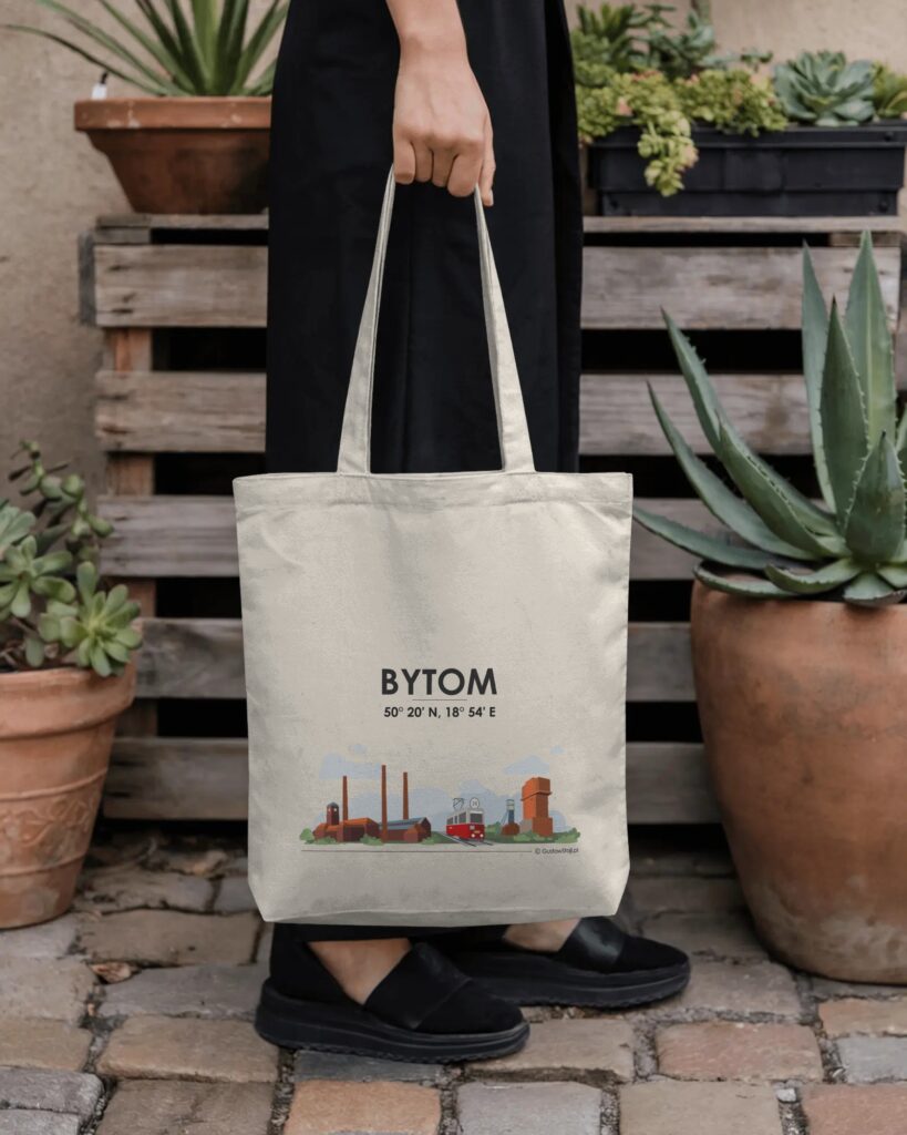 Osoba w czarnym stroju trzyma w ręku kremową torbę materiałową na tle drewnianych palet i roślin doniczkowych. Na torbie widnieje czarny napis „BYTOM”, pod nim współrzędne geograficzne oraz kolorowa ilustracja przedstawiająca Elektrociepłownię Szombierki i czerwony tramwaj.