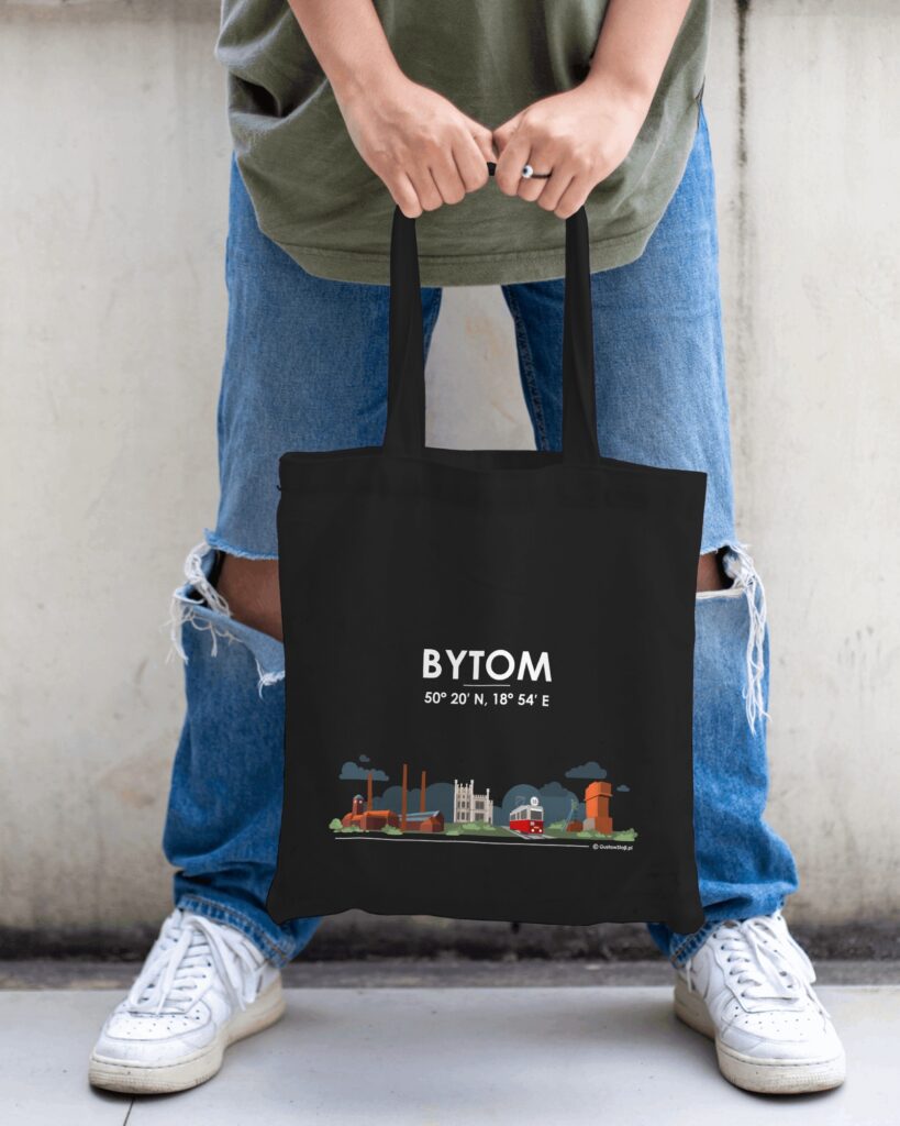 Osoba w dżinsach z przetarciami trzyma w dłoniach czarną torbę materiałową z białym napisem „BYTOM”, współrzędnymi geograficznymi oraz grafiką prezentującą panoramę architektury Bytomia i czerwony tramwaj nr 38.