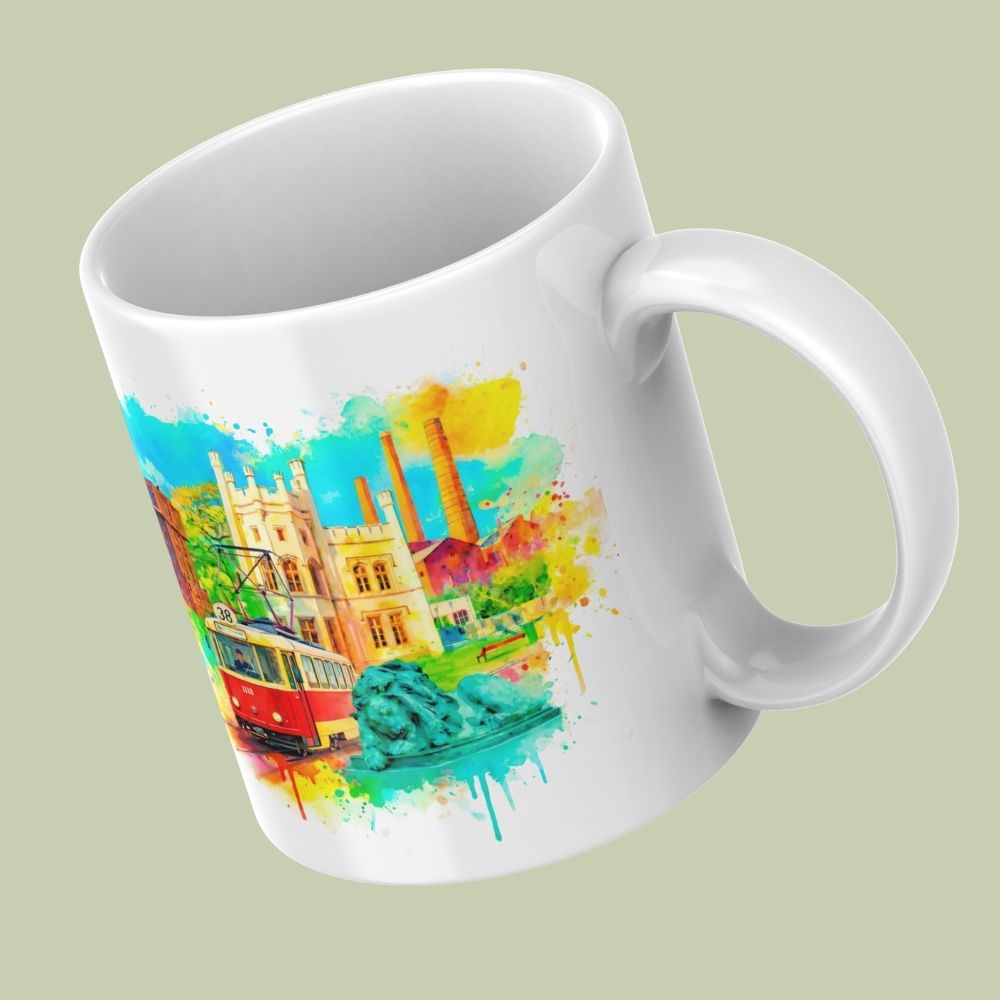 Biały kubek ceramiczny zaprezentowany pod kątem na jasnoszałwiowym tle. Na kubku wydrukowana jest kolorowa, akwarelowa grafika przedstawiająca symbole Bytomia: czerwony tramwaj linii 38, rzeźbę lwa oraz architekturę miejską i fabryczne kominy w otoczeniu barwnych plam farby.