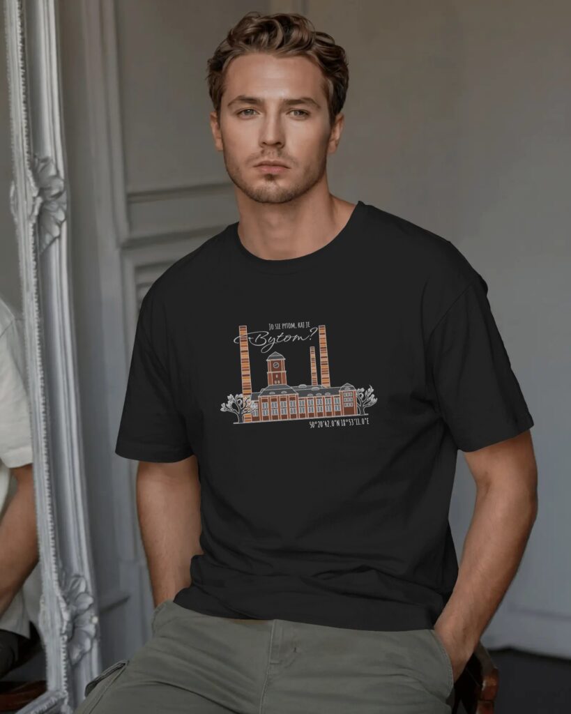 Zdjęcie portretowe mężczyzny stojącego przodem, w czarnym t-shircie. Na koszulce widnieje biało-pomarańczowa grafika przedstawiająca Elektrociepłownię Szombierki z napisem w języku śląskim „JO SIĘ PYTOM, KAJ JE BYTOM?” oraz współrzędnymi geograficznymi. Mężczyzna ma ręce w kieszeniach jasnych spodni, w tle widać fragment ozdobnej, białej ramy lustra.