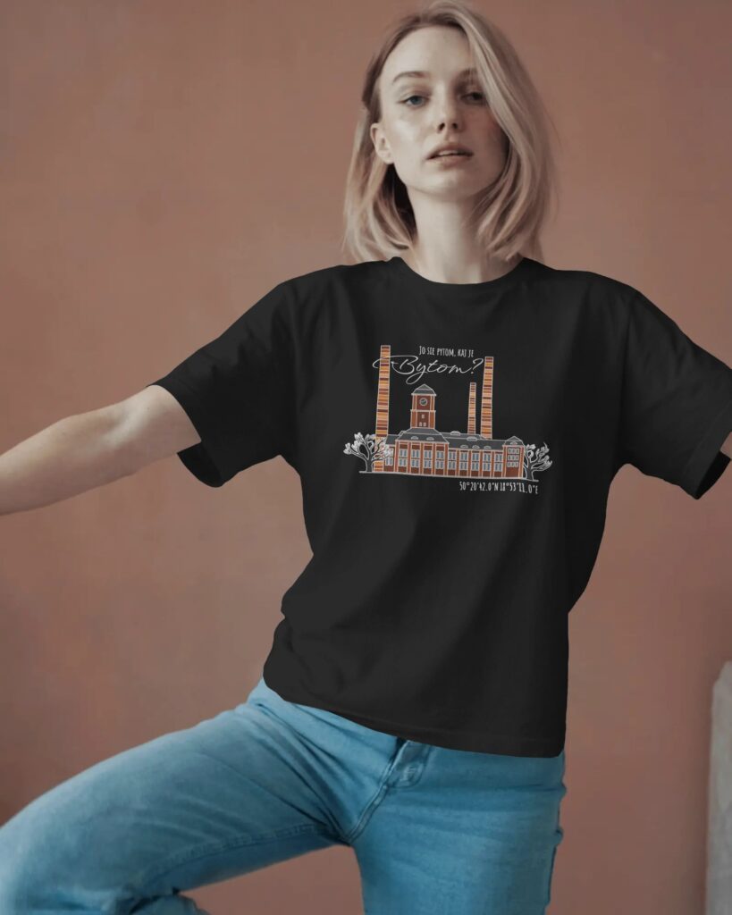 Zdjęcie kobiety w dynamicznej pozie, stojącej na jednej nodze, ubranej w czarny t-shirt. Na koszulce widoczna jest grafika z Elektrociepłownią Szombierki i napisem po śląsku „JO SIĘ PYTOM, KAJ JE BYTOM?”. Kobieta patrzy w bok, tło jest jednolite, w ciepłym, brązowym odcieniu.