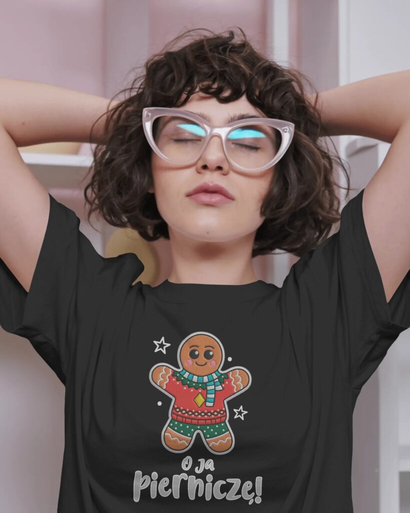 Kobieta o ciemnych, kręconych włosach i w jasnych okularach o ostrych oprawkach, trzymająca dłonie za głową. Ma na sobie czarny T-shirt oversize z nadrukiem przedstawiającym ilustrację piernikowego ludzika ubranego w świąteczny sweter oraz napis „O ja Pierniczę!” umieszczony tuż pod grafiką.