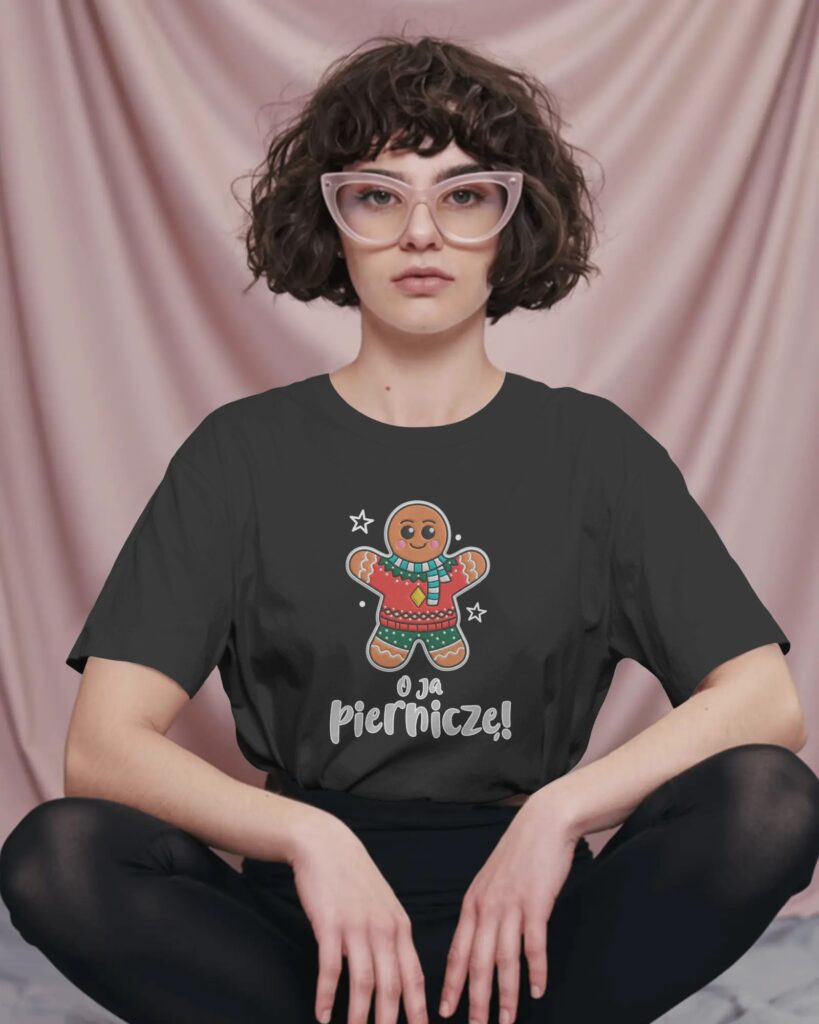 Kobieta z krótkimi, kręconymi włosami w jasnych okularach, siedząca ze skrzyżowanymi nogami na tle podwieszonej, różowej tkaniny. Ubrana jest w czarny T-shirt oraz czarne getry. Na środku koszulki widnieje ilustracja uśmiechniętego piernikowego ludzika w zimowym swetrze, z umieszczonym poniżej białym napisem „O ja Pierniczę!”.