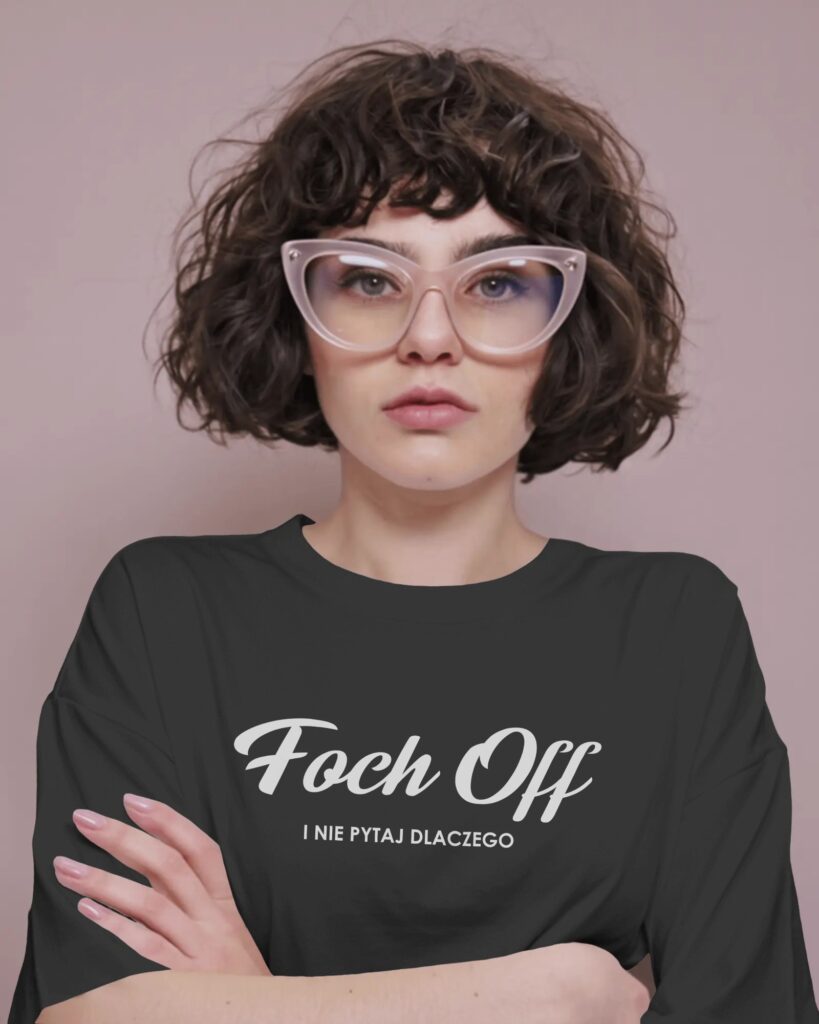 Kobieta o ciemnych, kręconych włosach do ramion i w jasnych okularach korekcyjnych w kształcie kocich oczu, stoi na stonowanym, różowawym tle z dłońmi skrzyżowanymi na piersi. Patrzy prosto w aparat. Nosi czarny t-shirt z białym, ozdobnym napisem „Foch Off” i dopełniającym tekstem „I NIE PYTAJ DLACZEGO”.