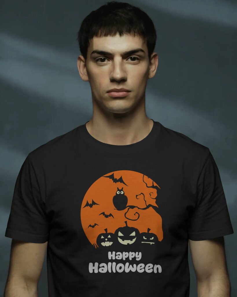 Młody mężczyzna o ciemnych, krótkich włosach i poważnym wyrazie twarzy, pozujący na nastrojowym, ciemnoszarym tle. Ubrany jest w czarny T-shirt z centralnie umieszczonym nadrukiem przedstawiającym pomarańczową pełnię księżyca, sylwetki nietoperzy, sowę, trzy dynie z wyciętymi twarzami oraz biały napis „Happy Halloween”.