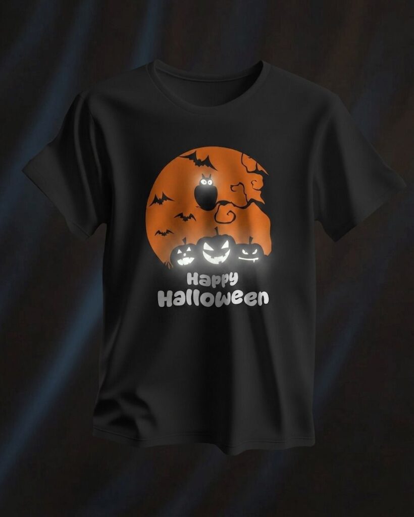 Czarny T-shirt zaprezentowany na ciemnym, eleganckim tle z delikatnymi załamaniami światła. Na środku koszulki znajduje się grafika przedstawiająca duży, pomarańczowy księżyc w pełni z sylwetkami nietoperzy i sowy na gałęzi. U podnóża księżyca widnieją trzy uśmiechnięte dynie halloweenowe z rozświetlonymi oczami i ustami, a pod nimi umieszczono biały napis „Happy Halloween”.