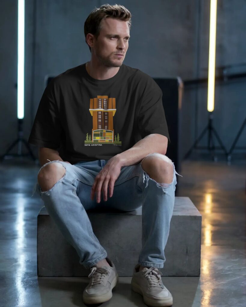 Mężczyzna siedzący na betonowym sześcianie w surowym studio fotograficznym z podłużnymi lampami w tle. Nosi czarny t-shirt z nadrukiem przedstawiającym wieżę wyciągową Szyb Krystyna oraz napis „SZYB KRYSTYNA”. Mężczyzna ma na sobie jasnoniebieskie jeansy z przetarciami na kolanach oraz beżowe buty sportowe.