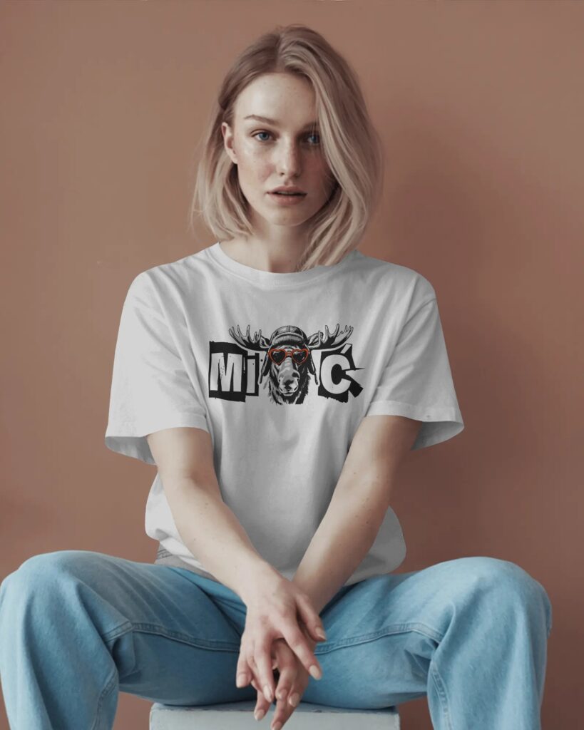 Kobieta o blond włosach siedząca na stołku ze skrzyżowanymi nogami na tle ciepłej, brązowej ściany. Ma na sobie białą koszulkę T-shirt o luźnym kroju oraz jasnoniebieskie dżinsy. Na piersi znajduje się nadruk przedstawiający napis „MIŁOŚĆ” z grafiką głowy łosia w czerwonych okularach-serduszkach i czapce pilotce.