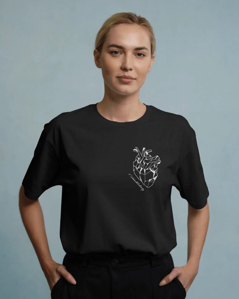 Zdjęcie portretowe młodej kobiety z jasnymi, spiętymi włosami, stojącej na jednolitym, jasnoniebieskim tle. Kobieta ma na sobie czarny t-shirt, na którym po lewej stronie klatki piersiowej znajduje się biały, geometryczny nadruk przedstawiający anatomiczne serce. Poniżej serca widać napis „Hercklekoty”. Kobieta trzyma ręce w kieszeniach czarnych spodni.