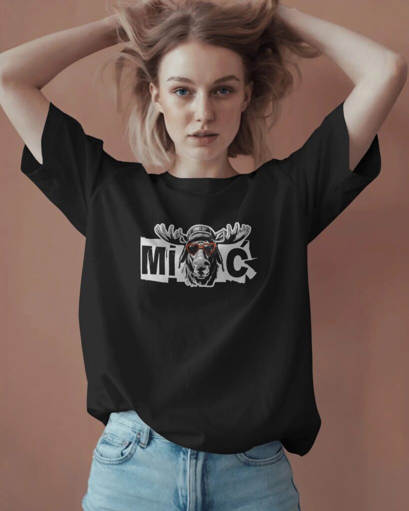 Młoda kobieta o jasnych włosach, trzymająca dłonie przy głowie, pozująca na ciepłym, brązowym tle. Ubrana jest w czarny T-shirt oversize oraz jasne dżinsy. Na klatce piersiowej widnieje graficzny napis „MIŁOŚĆ” z wizerunkiem łosia w czapce pilotce i czerwonych okularach w kształcie serc.