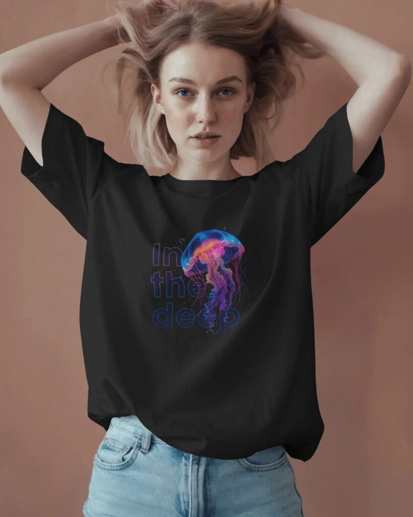 Jasnowłosa kobieta z dłońmi we włosach, uniesionymi nad głową, pozująca na gładkim, brązowym tle. Ma na sobie czarny T-shirt oversize oraz jasnoniebieskie dżinsy. Na piersi koszulki widnieje neonowa grafika przedstawiająca świecącą meduzę w odcieniach różu, fioletu i niebieskiego z wkomponowanym w tło napisem „In the deep”.