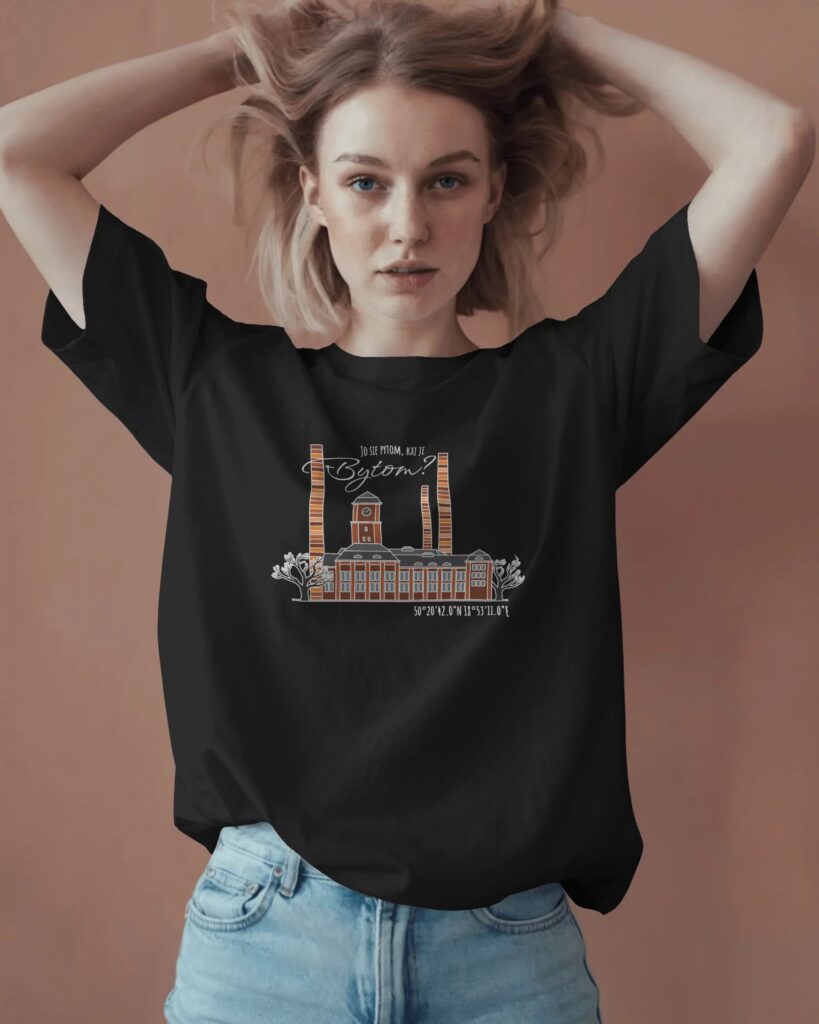 Zdjęcie młodej kobiety z blond włosami, pozującej w czarnym t-shircie. Kobieta trzyma ręce uniesione w górze, mierzwiąc włosy, i patrzy w obiektyw. Na środku koszulki znajduje się nadruk przedstawiający zabytkową Elektrociepłownię Szombierki oraz napis: „JO SIĘ PYTOM, KAJ JE BYTOM?” wraz ze współrzędnymi. Nosi jasne jeansy, tło jest gładkie, w kolorze terakoty.