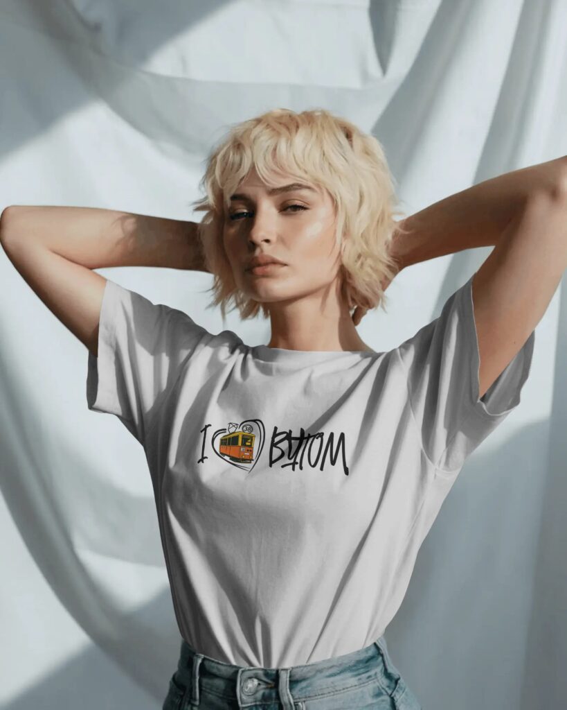 Młoda kobieta o krótkich, blond włosach z grzywką, pozująca z dłońmi za głową na tle marszczonej, białej tkaniny. Ma na sobie jasnoszary T-shirt wpuszczony w niebieskie dżinsy. Na piersi koszulki znajduje się czarny nadruk „I [serce] BYTOM” z ilustracją klasycznego, zabytkowego tramwaju linii 38 wpisanego w zarys serca.